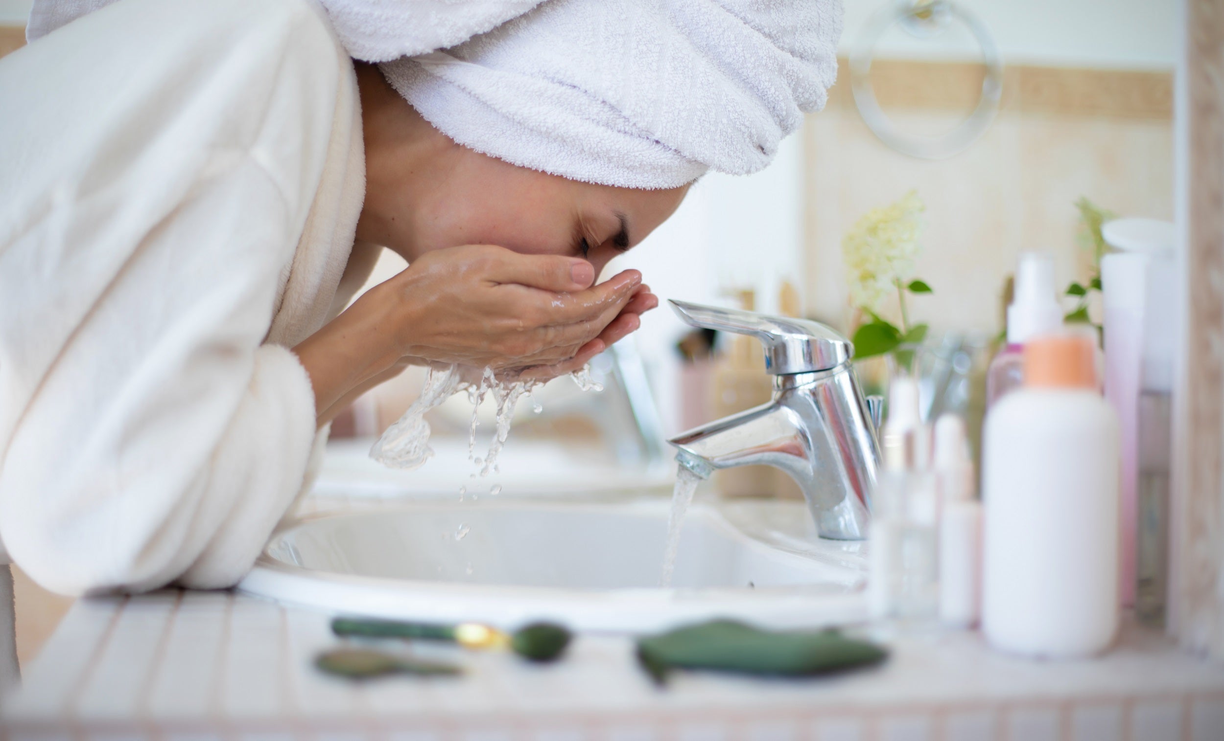 A woman in a towel and bathrobe washes her face at a bathroom sink